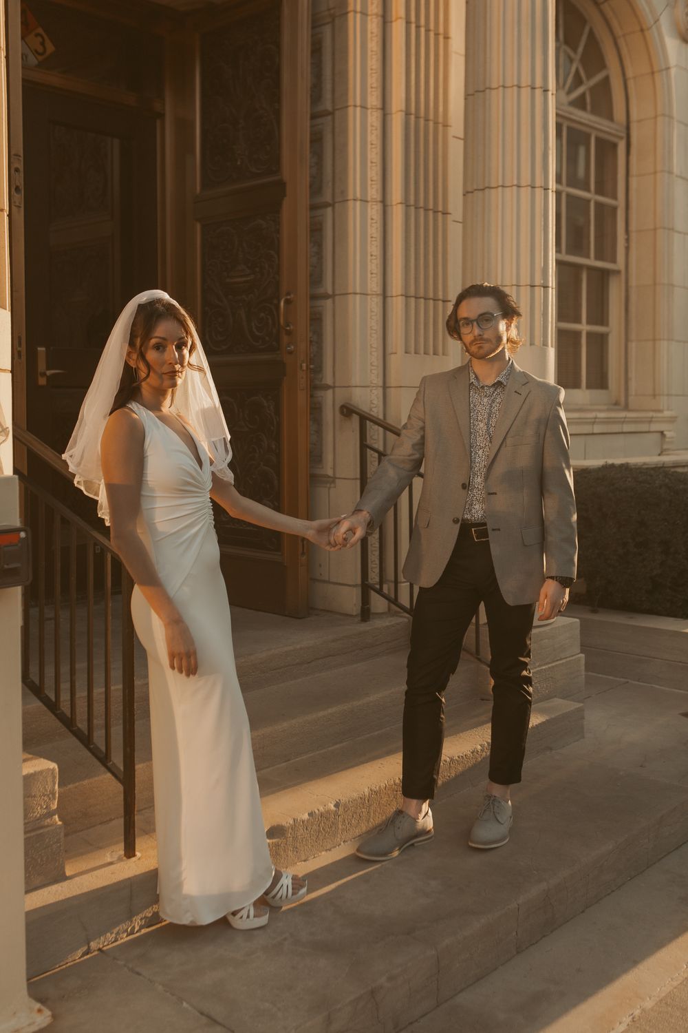 A wedding couple standing on stone steps outside a historic building with elegant columns in warm golden light.