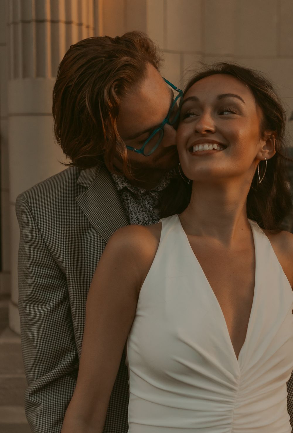 A romantic intimate moment captured between a couple in formal attire during golden hour lighting against a columned backdrop.