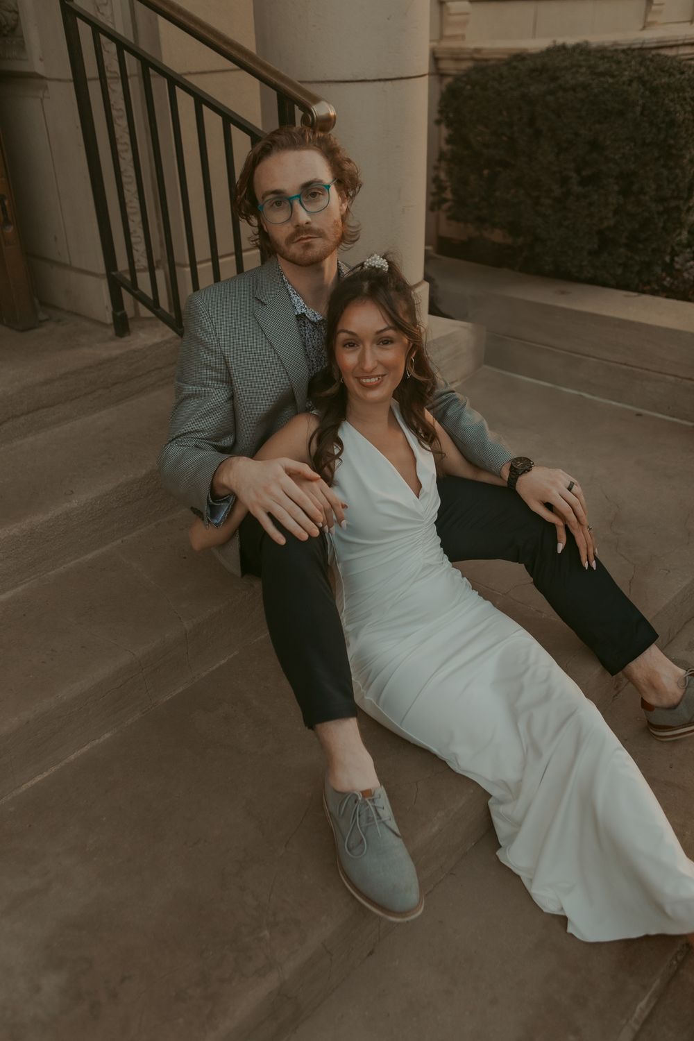A couple in formal attire sitting on outdoor steps, with the woman wearing a white dress and the man in a gray suit.