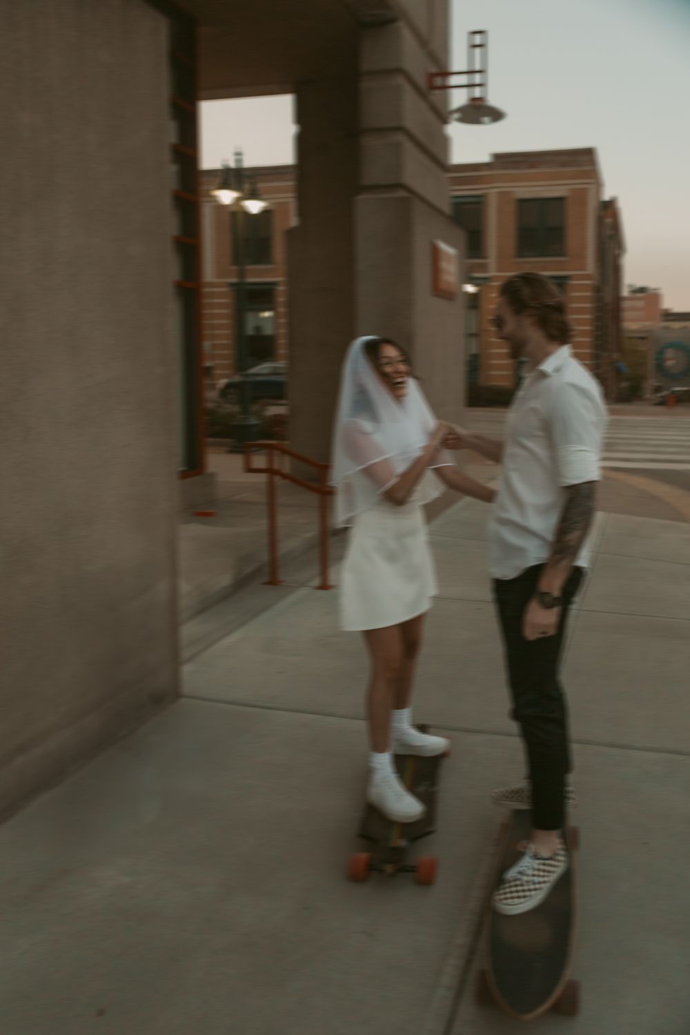 Two people on skateboards stand talking on a city sidewalk at dusk.