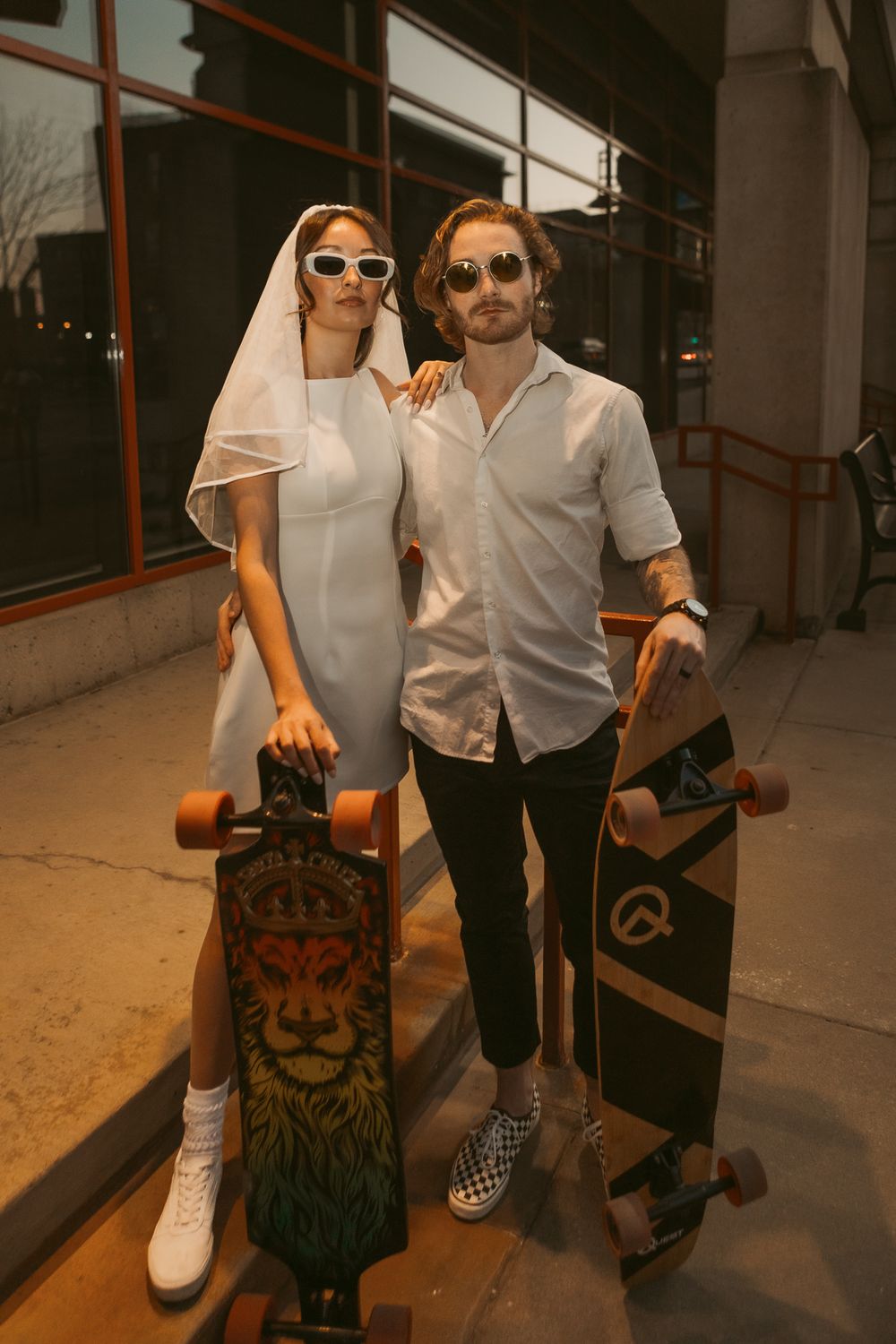A stylish couple in sunglasses poses with their longboards outside a building in a warm sunset atmosphere.
