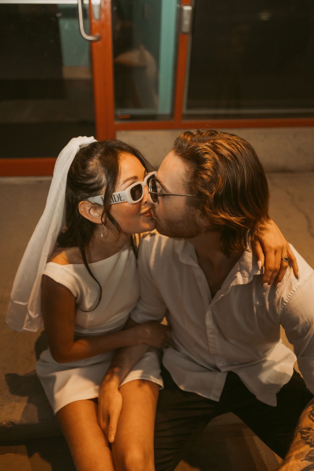 A romantic couple in white outfits share a sweet kiss while sitting together on the floor at sunset.