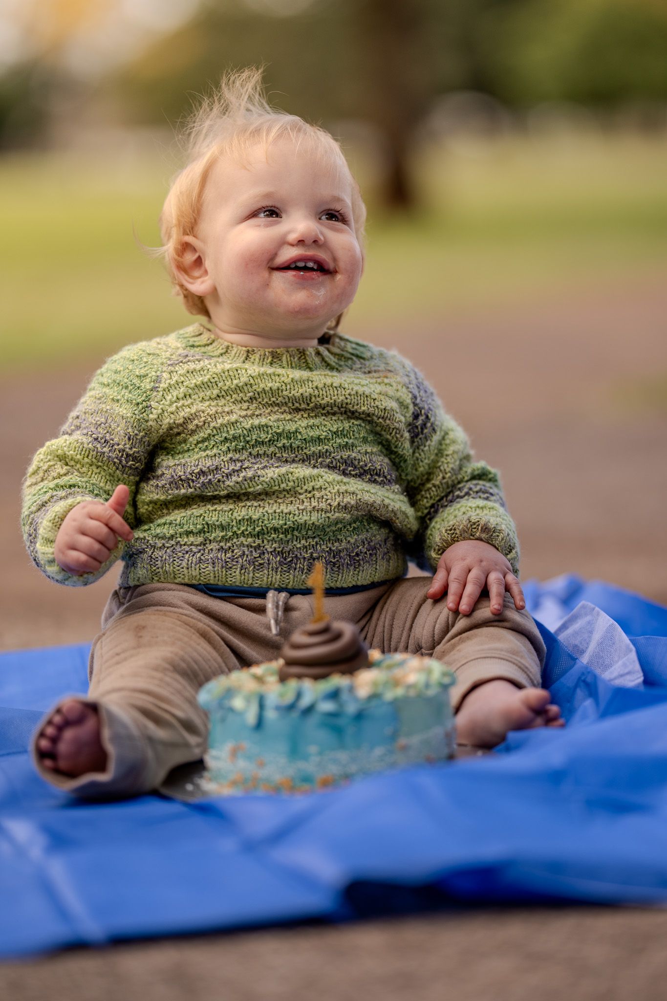 Baby wearing green knit sweater and brown pants sits on blue blanket with birthday cake smiling with joy.