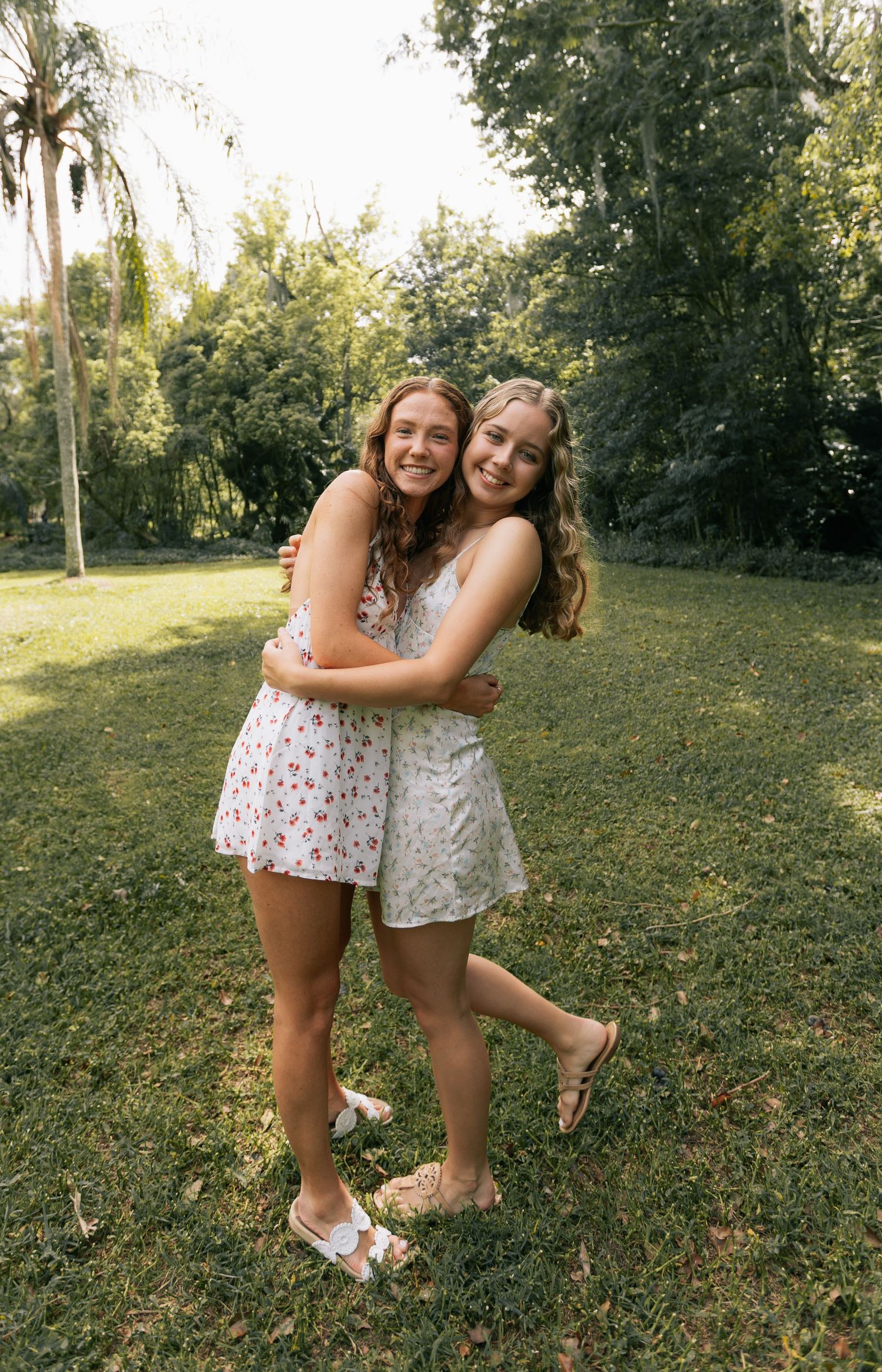 Two smiling young women in floral sundresses hug on a sunny grassy lawn surrounded by green trees.