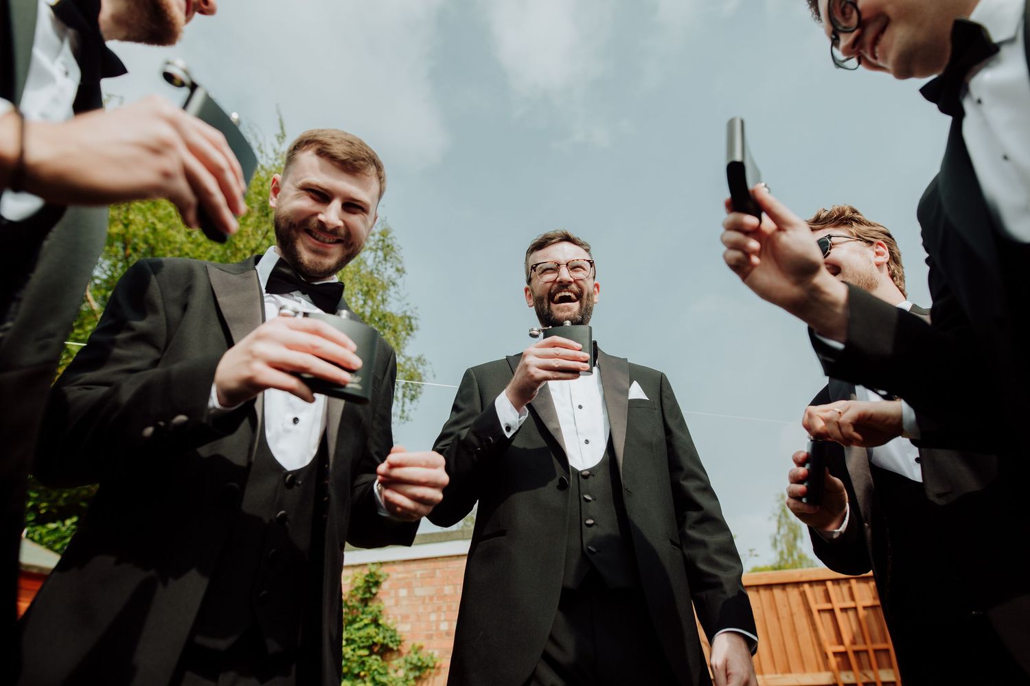 People in formal black tuxedos and bow ties laughing and holding beverages at an outdoor celebration.