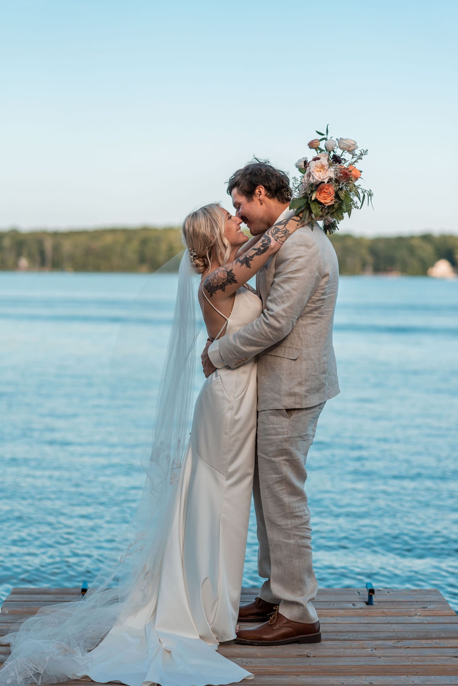 A romantic wedding couple shares an intimate moment on a wooden dock overlooking a serene lake at sunset.