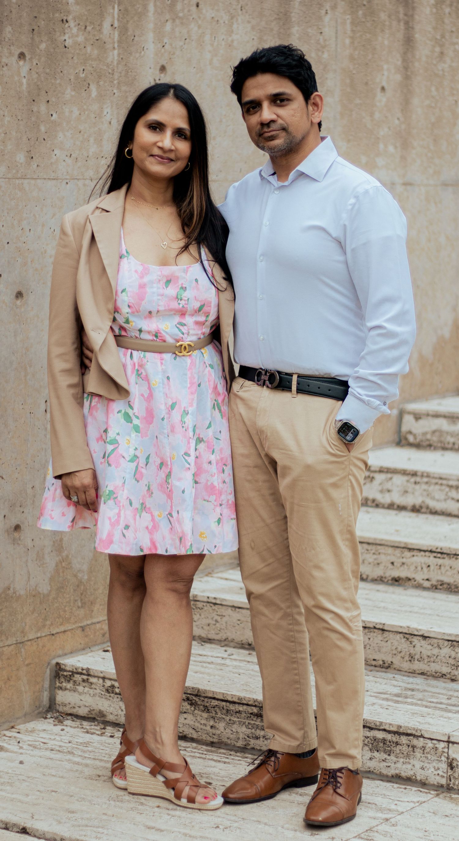A couple poses on stone steps, wearing coordinated casual outfits with her in a floral dress and him in khakis and white shirt.