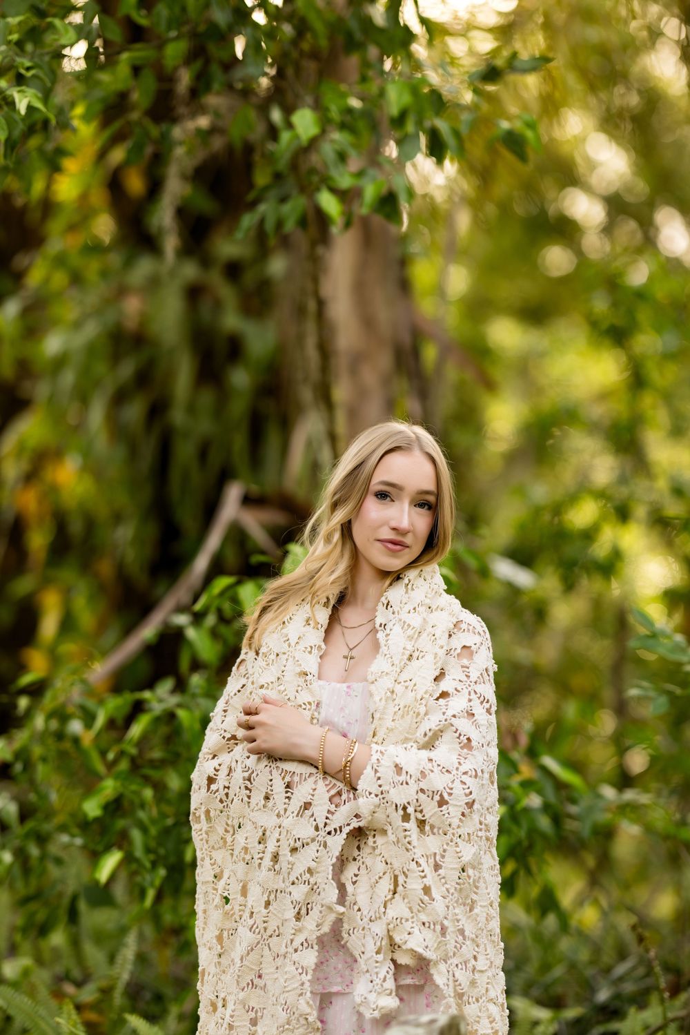 Figure wrapped in an elegant cream lace shawl poses against a verdant garden backdrop.