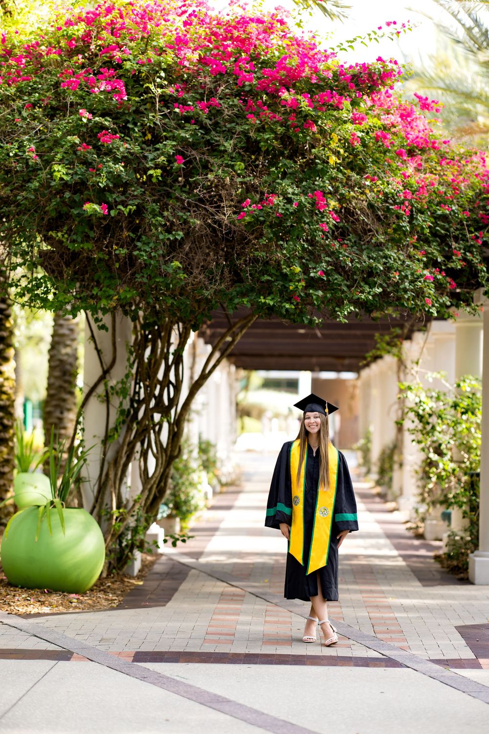 A smiling graduate in black cap and gown with yellow stole walks under a flowering bougainvillea pergola.