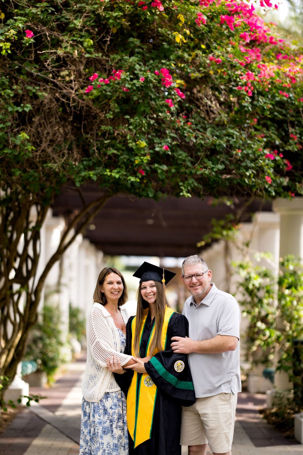 A graduate in cap and gown poses with her proud parents under a flowering bougainvillea archway.