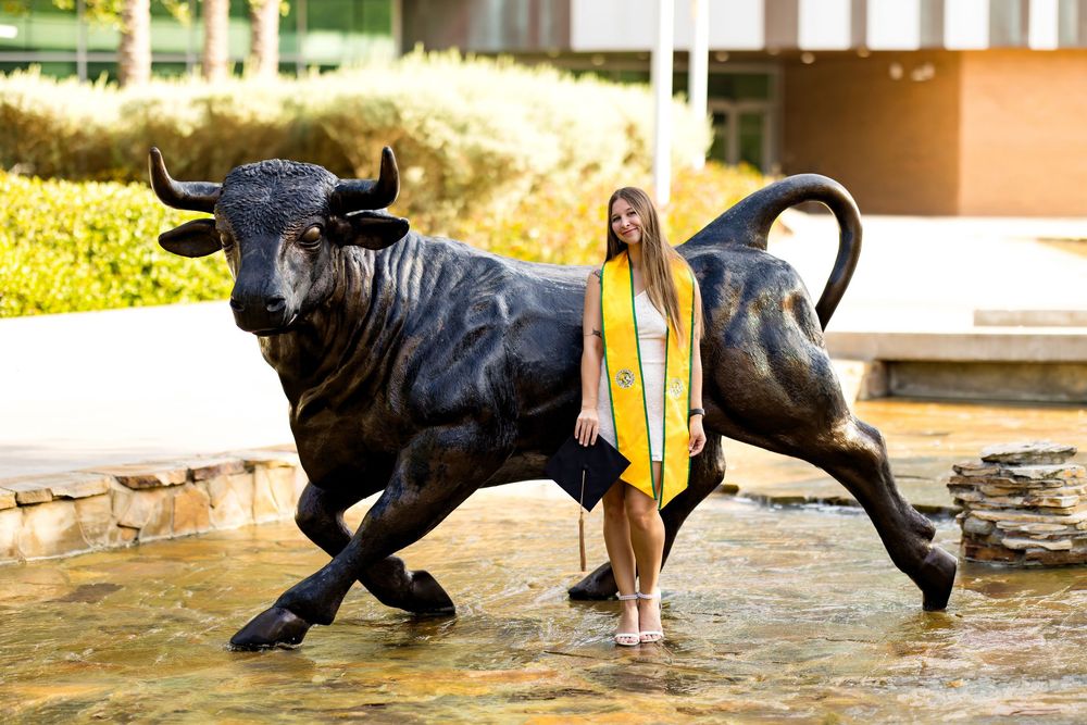 Graduate in cap and gown poses beside a bronze bull statue in a fountain, wearing a gold sash.