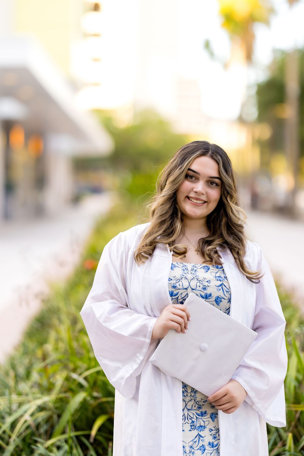 Graduate holding diploma in white graduation robe and blue floral dress by landscaped path.
