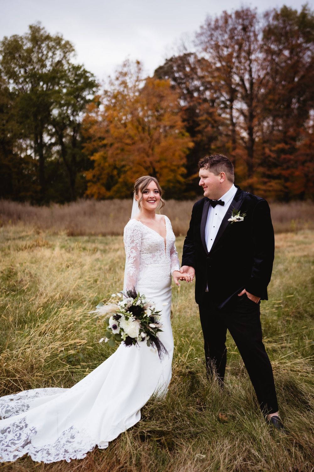 A bride and groom share candid moments in a rustic autumn field with tall grass and fall foliage in the background.