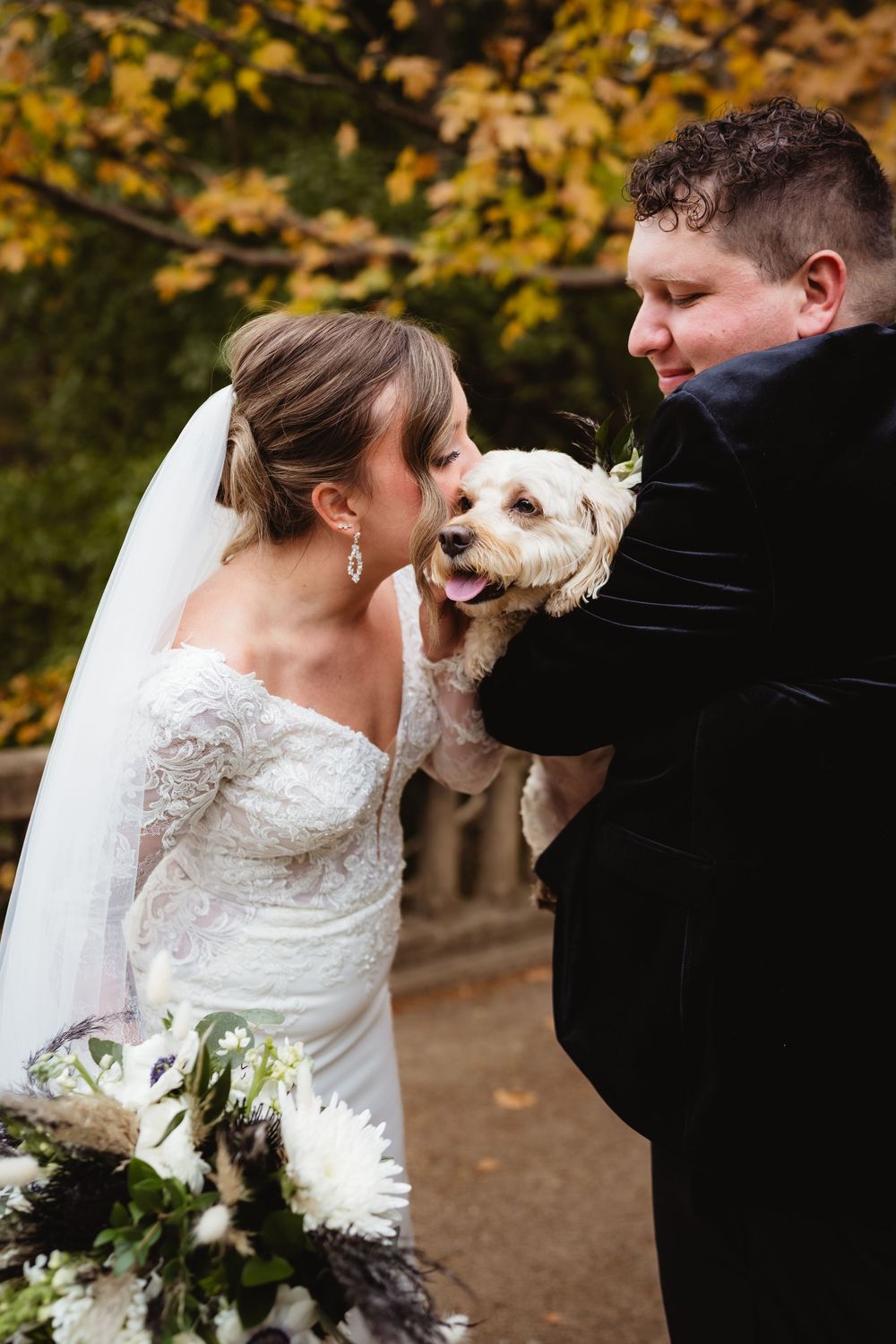 A couple in wedding attire shares a sweet moment with their small white dog during an autumn outdoor photoshoot.