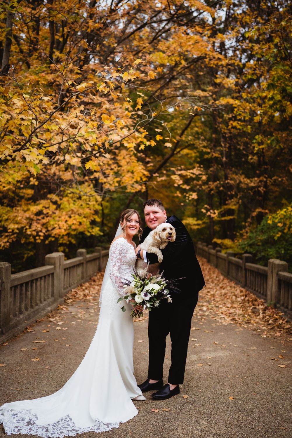 A couple in wedding attire stands on a bridge path surrounded by vibrant autumn foliage in golden hues.
