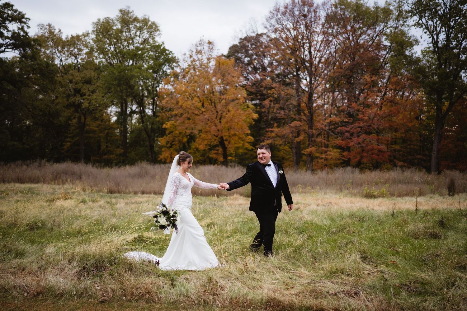 A couple in wedding attire dances together in a rustic autumn field with golden trees in the background.