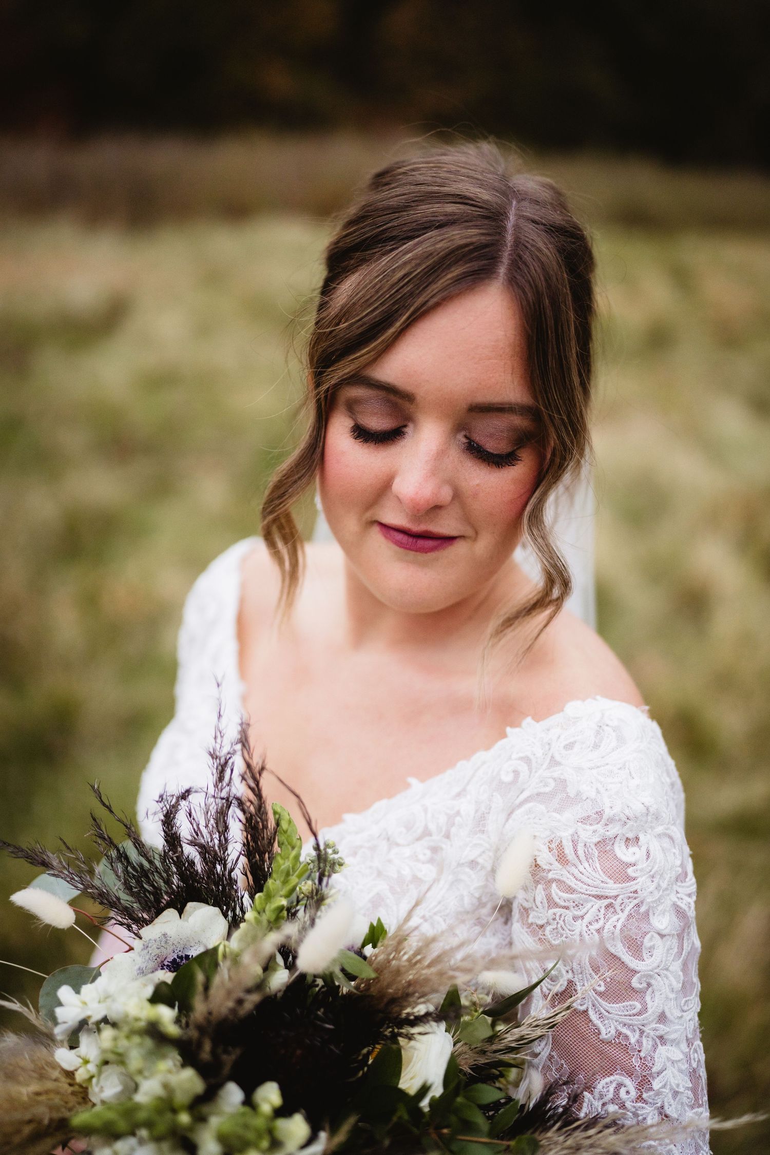 Bride in lace off-shoulder wedding dress looks down at rustic bouquet with white flowers and dark foliage.