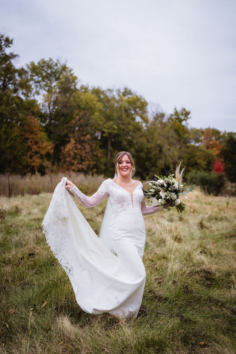 A bride in a flowing white lace wedding dress twirls gracefully in a rustic field surrounded by autumn trees.