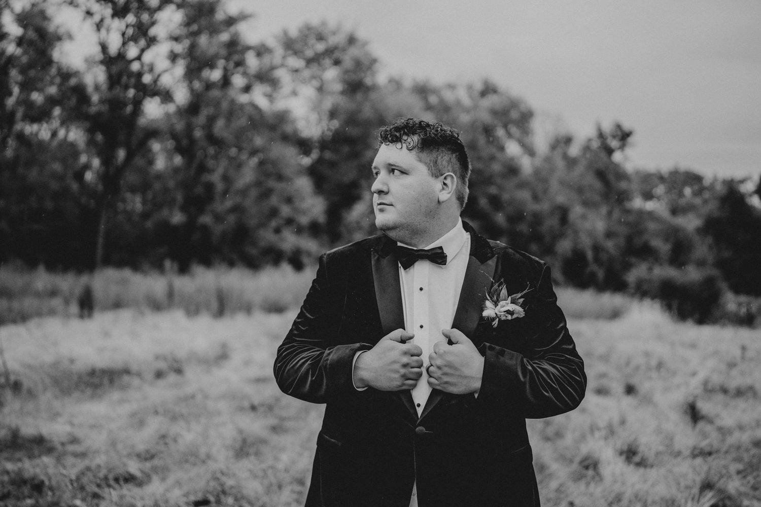 Black and white photo of a groom in a tuxedo adjusting his bowtie while standing in an outdoor field.