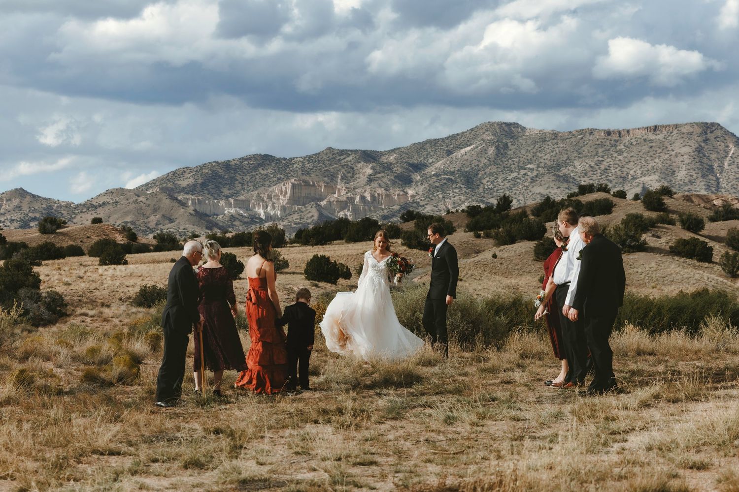 Outdoor wedding ceremony in a desert landscape with mountains, guests gathered around bride and groom under cloudy skies.