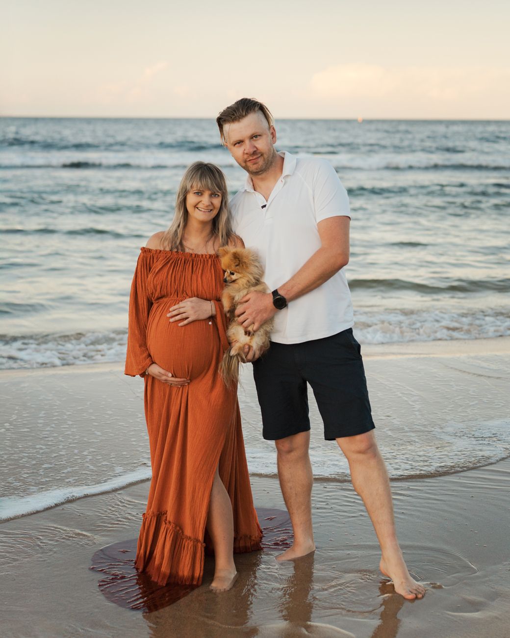 An expacting couple stands together on a sandy beach at sunset with their dog during a maternity photoshoot in Sydney Maroubra Beach