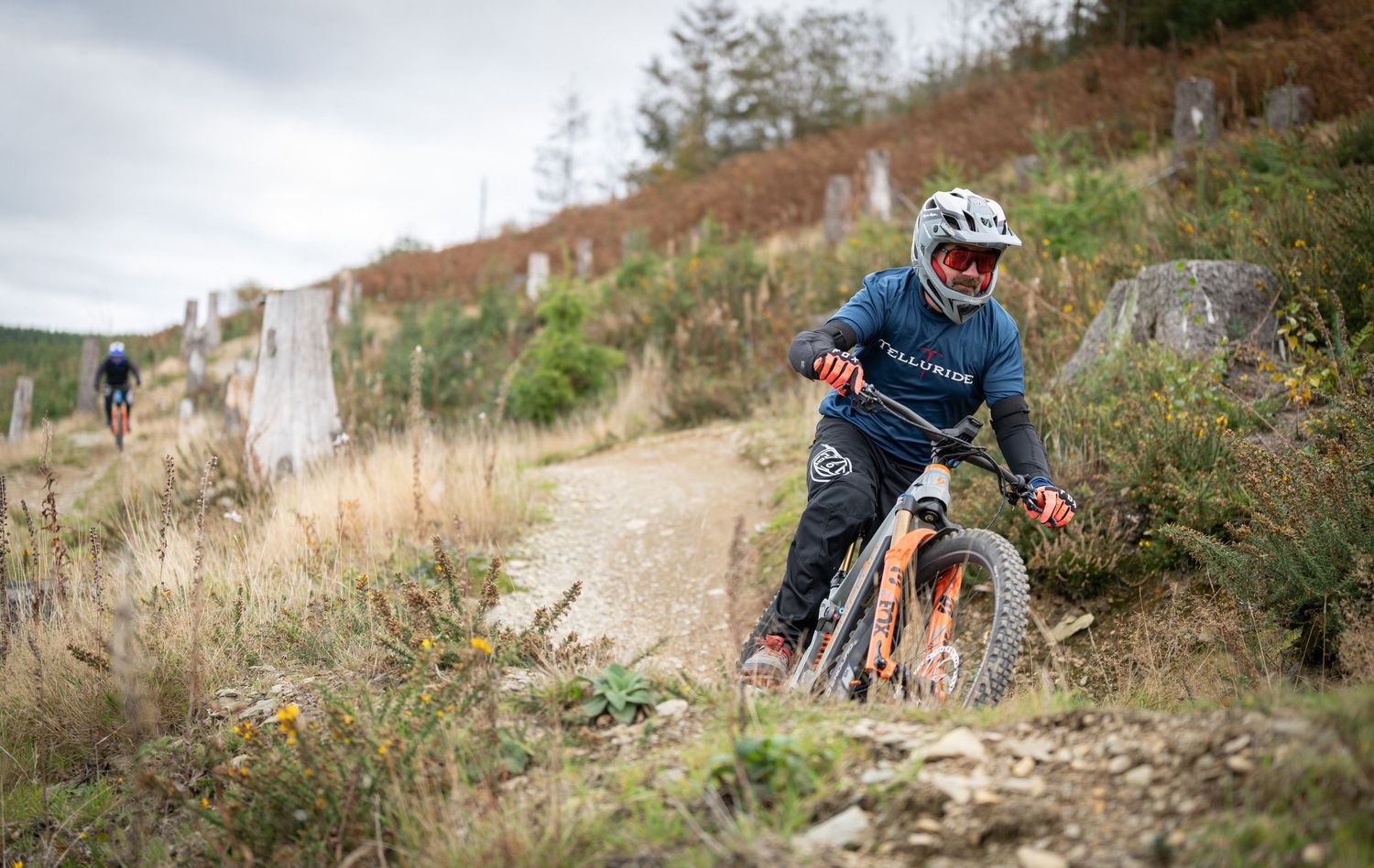 Mountain biker riding an orange bike on a dirt trail through a forest, wearing a helmet and blue jersey.