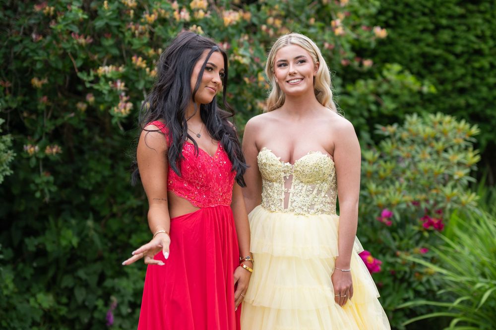Friends in elegant prom dresses pose together against a lush green garden backdrop.