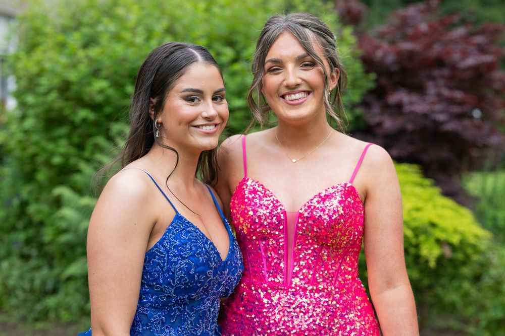 Friends in sparkling blue and pink formal dresses smile together in a garden setting.