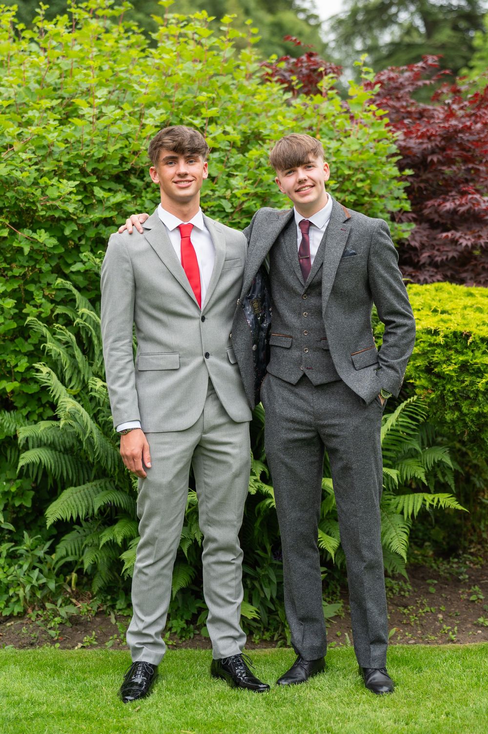 Two friends in formal suits, one grey and one charcoal, pose together on manicured garden grounds.