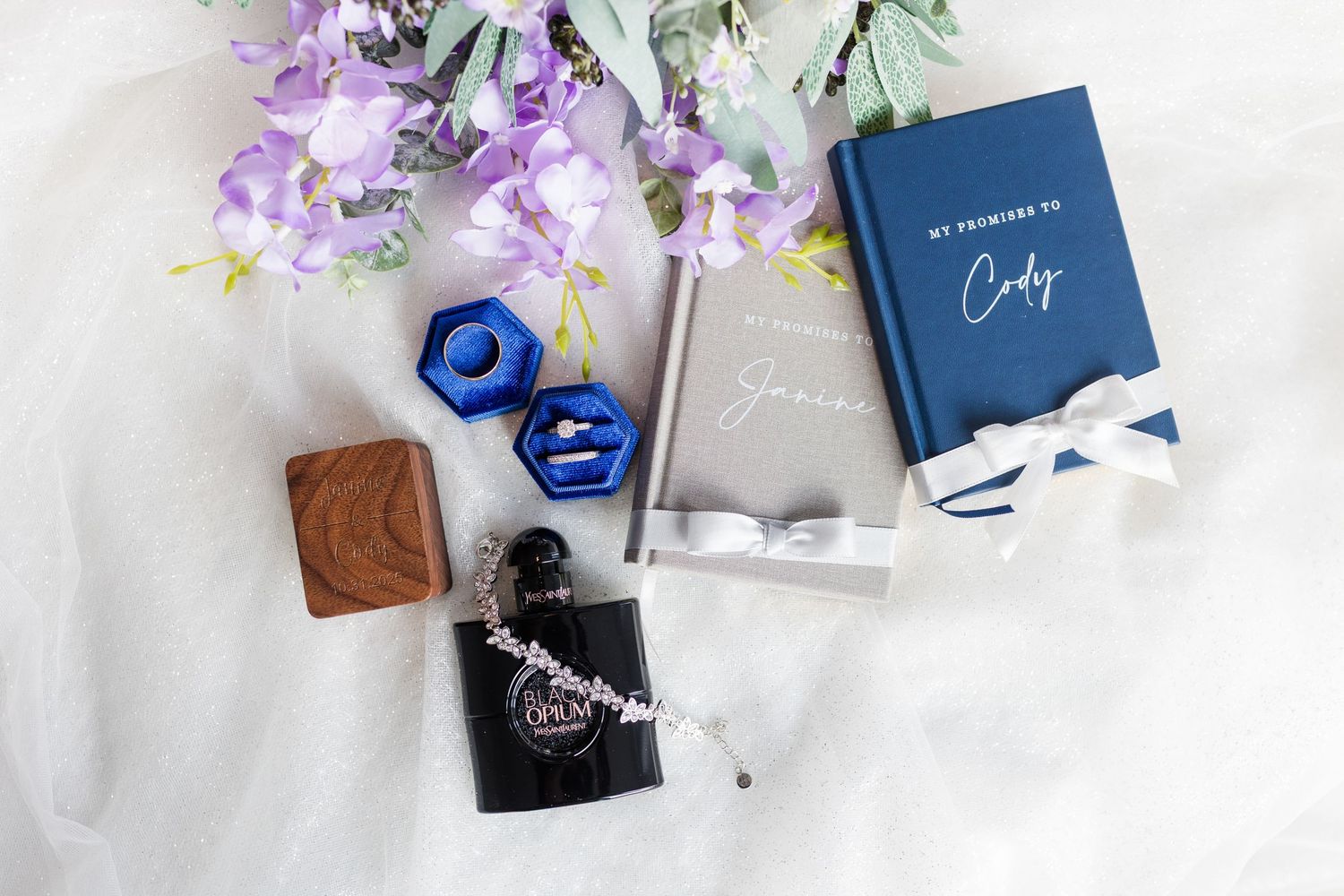 Luxury jewelry and beauty products arranged with purple flowers and navy blue boxes on white marble surface.