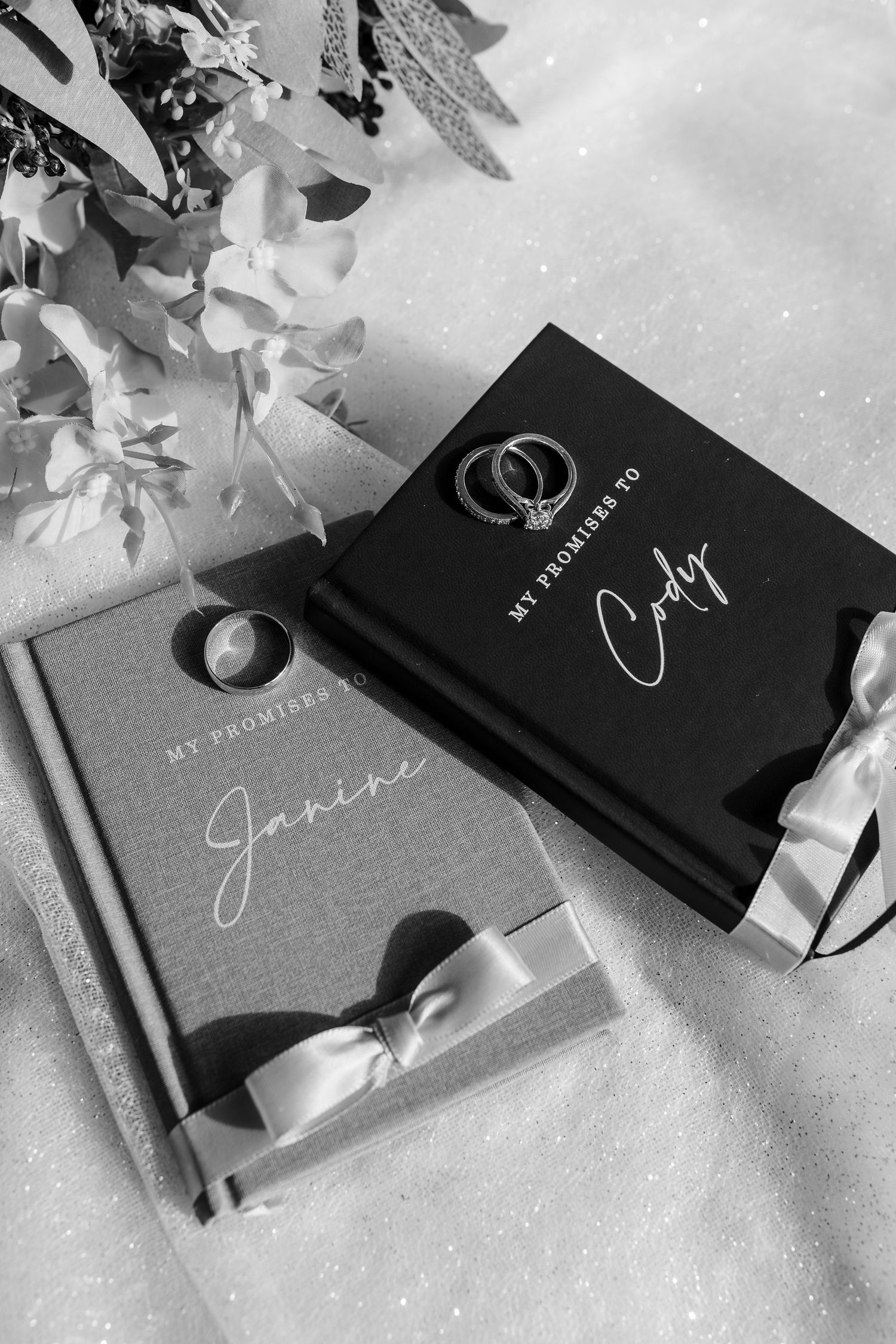 Black and white photo of elegant wedding vow books with calligraphy text arranged on a light surface with floral accents.
