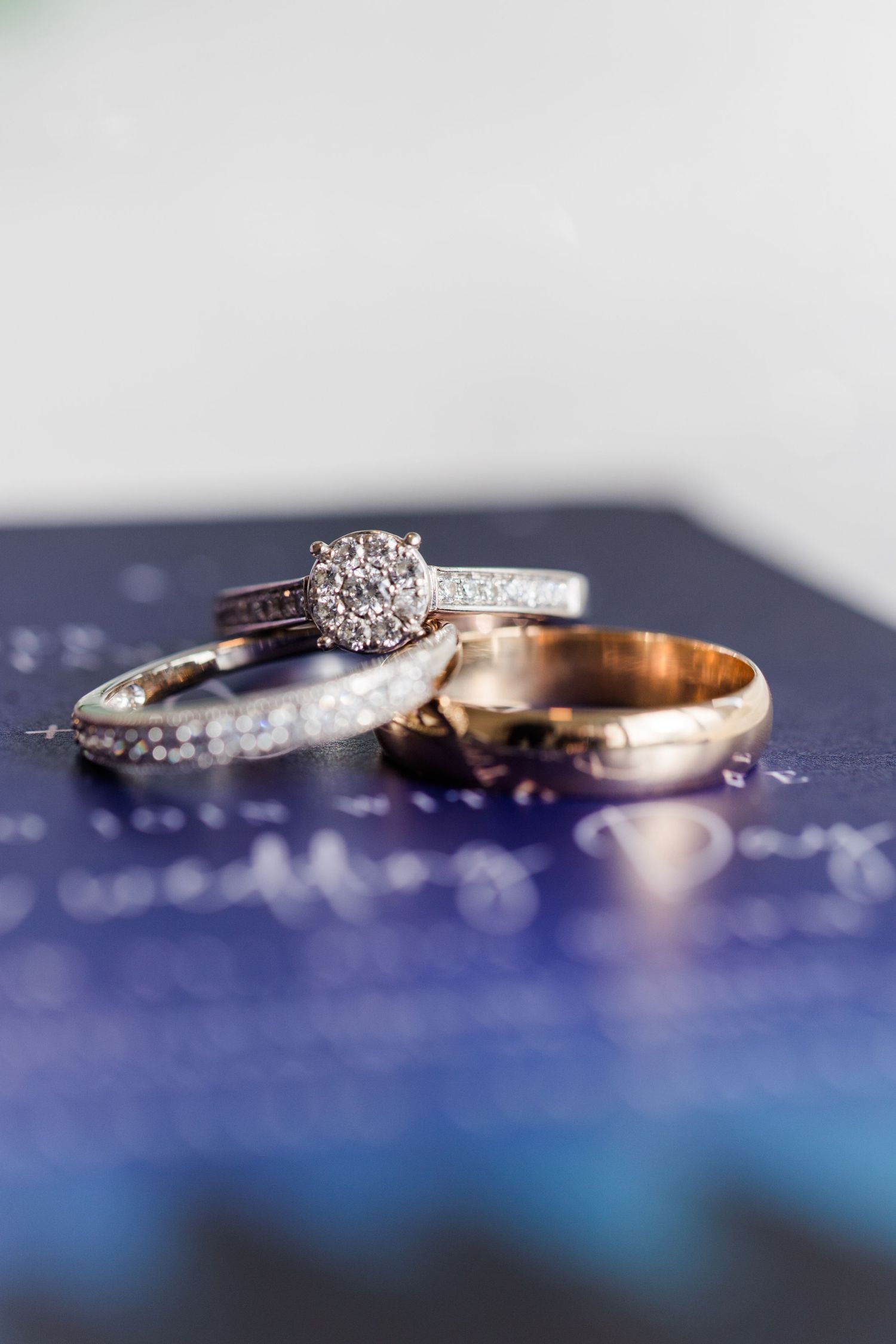 Close up of diamond engagement ring and wedding bands arranged on a blue surface with sparkling reflections.