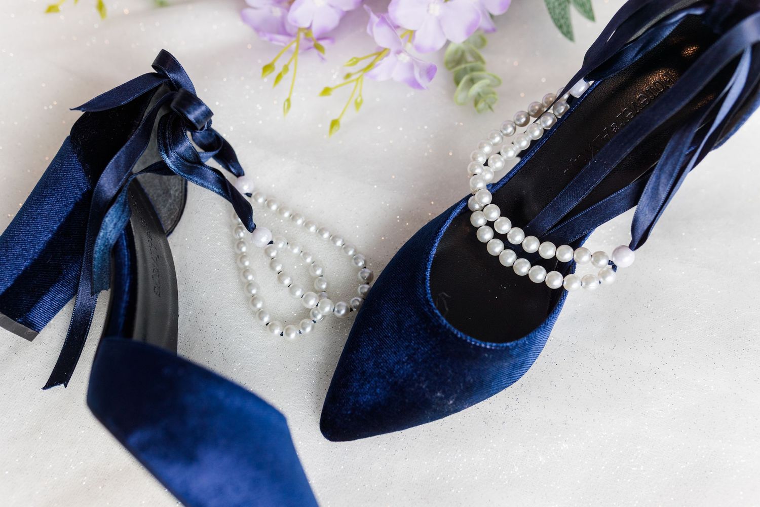 Navy blue velvet heels with pearl accessories and ribbon bows displayed on white lace fabric.