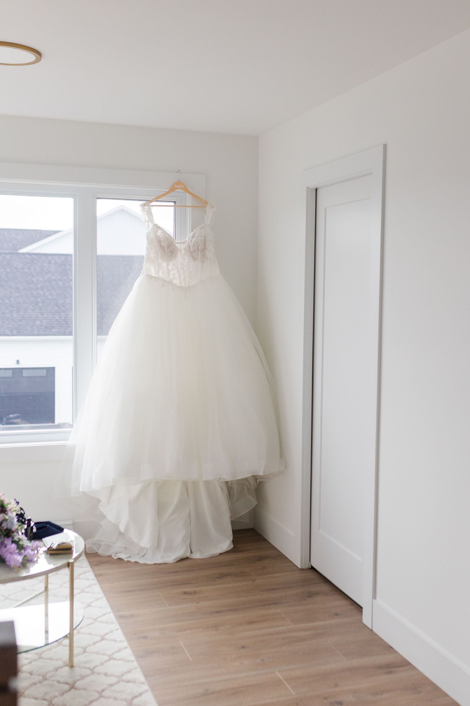 White wedding dress displayed on a hanger in a bright, minimalist room with light wooden flooring and white walls.