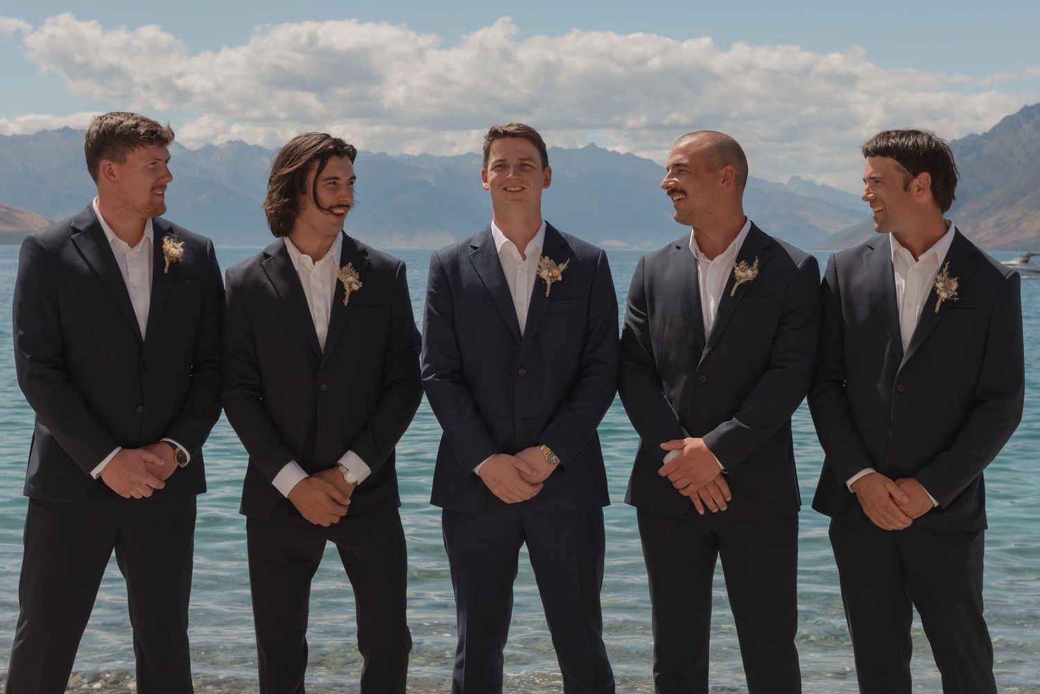 Groomsmen in black suits stand in a line posing for a wedding photo against a scenic mountain lake backdrop.