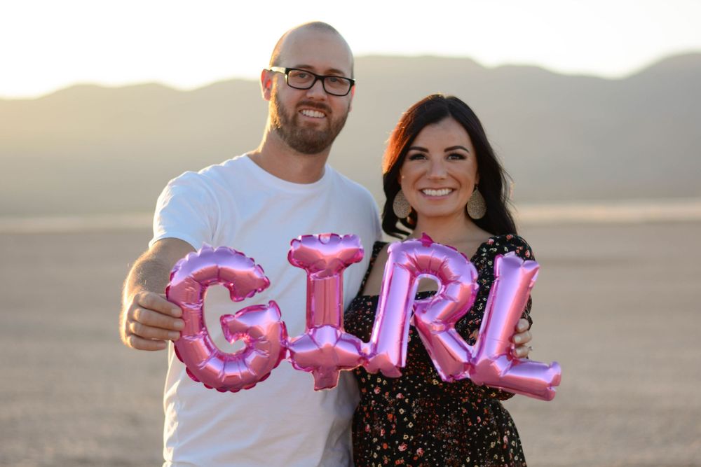 A group of people pose outdoors holding pink letter balloons spelling out GIRL during a gender reveal celebration.