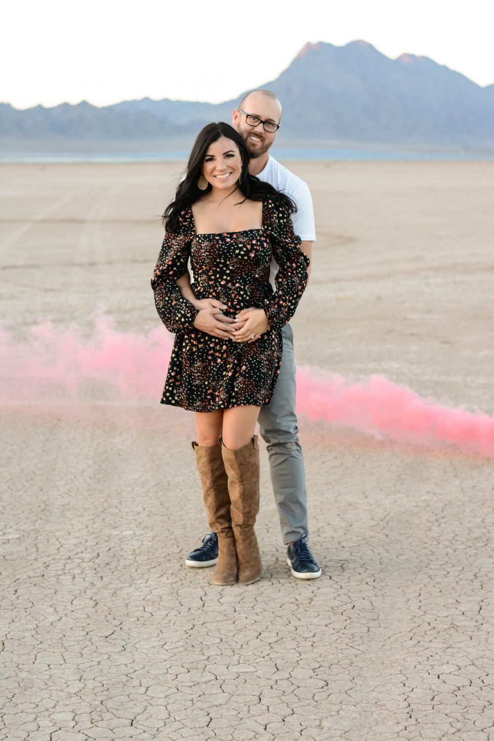 Couple embraces on cracked desert ground with pink smoke in the background during a romantic photoshoot.