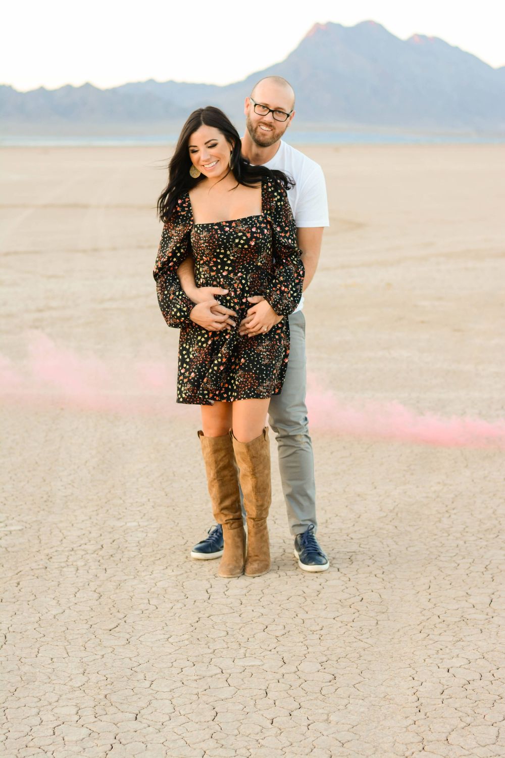 A couple embraces while standing on a cracked desert floor with mountains visible in the background.