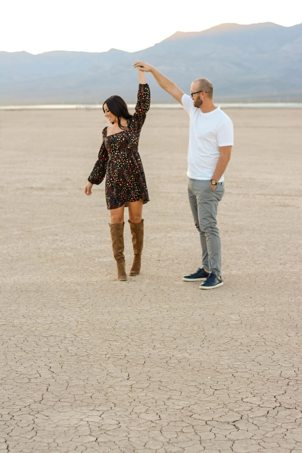 Two people dance and pose playfully on cracked desert ground with mountains visible in the background.