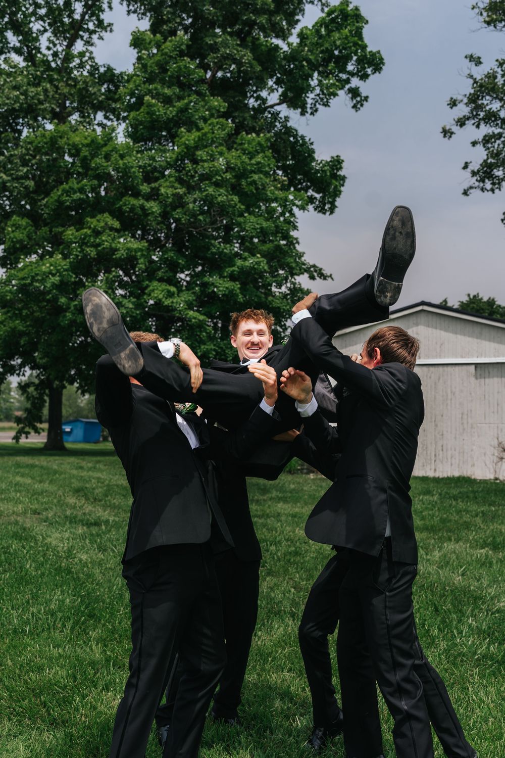 Two people in black suits playfully dance and dip together on a grassy field with trees in the background.