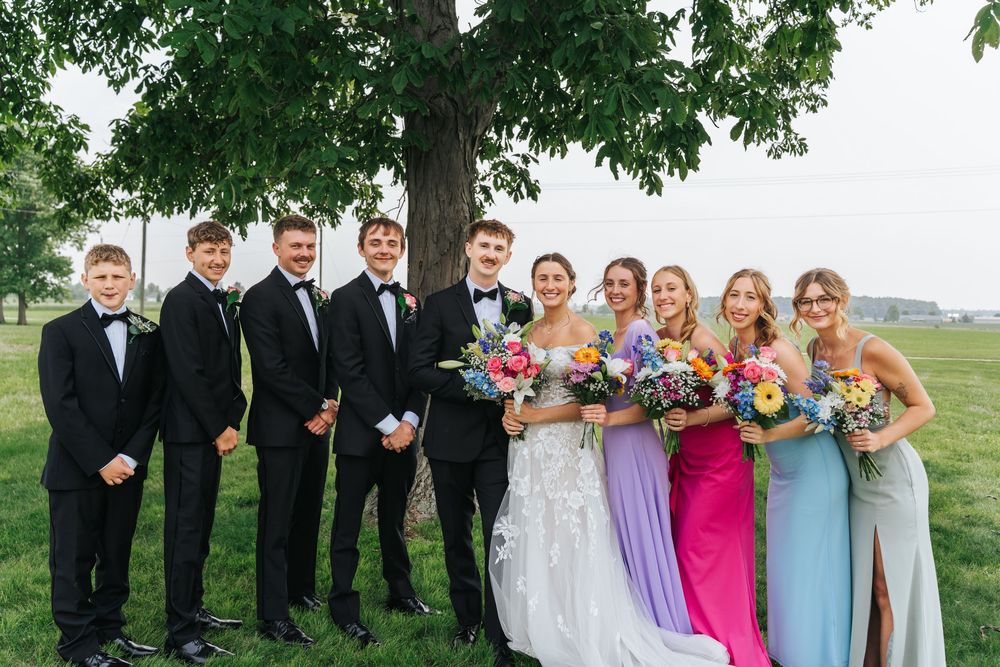 A wedding party stands together outdoors in formal attire with bridesmaids in colorful dresses and groomsmen in black suits.