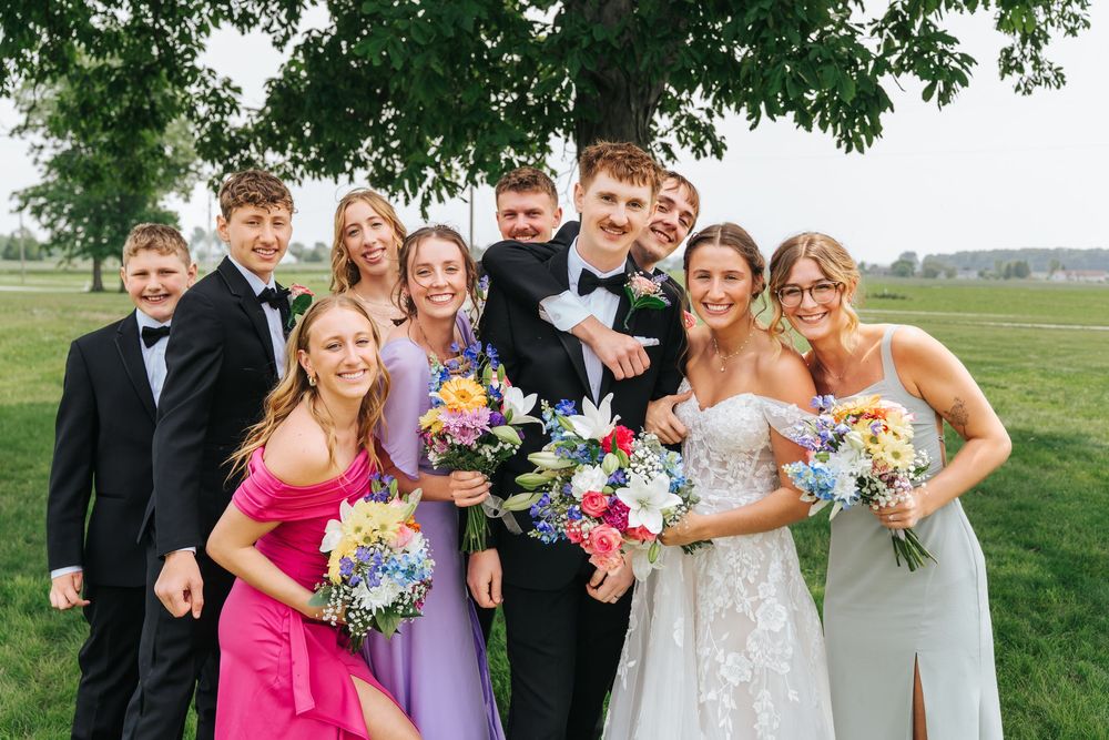 Wedding party in formal attire poses outdoors with colorful bouquets under a green tree on grassy field.