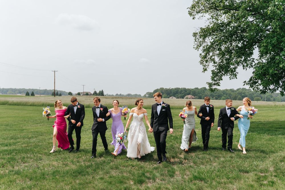 Large wedding party poses for formal group photos in an open grassy field on a summer day.