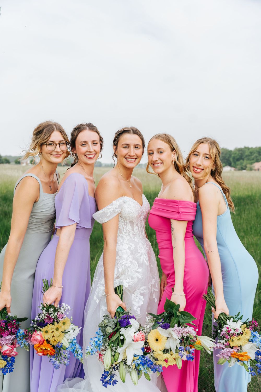 A bridal party wearing pastel dresses holds colorful bouquets while posing together in a sunny field.