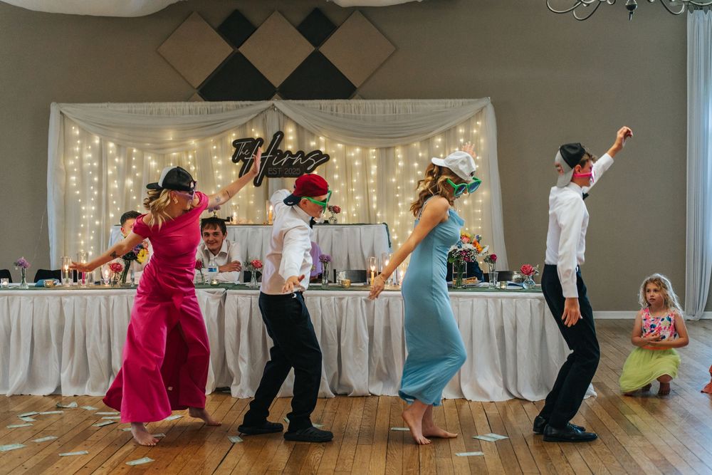 Wedding party members perform a choreographed dance sequence in a reception hall with twinkling lights in the background.