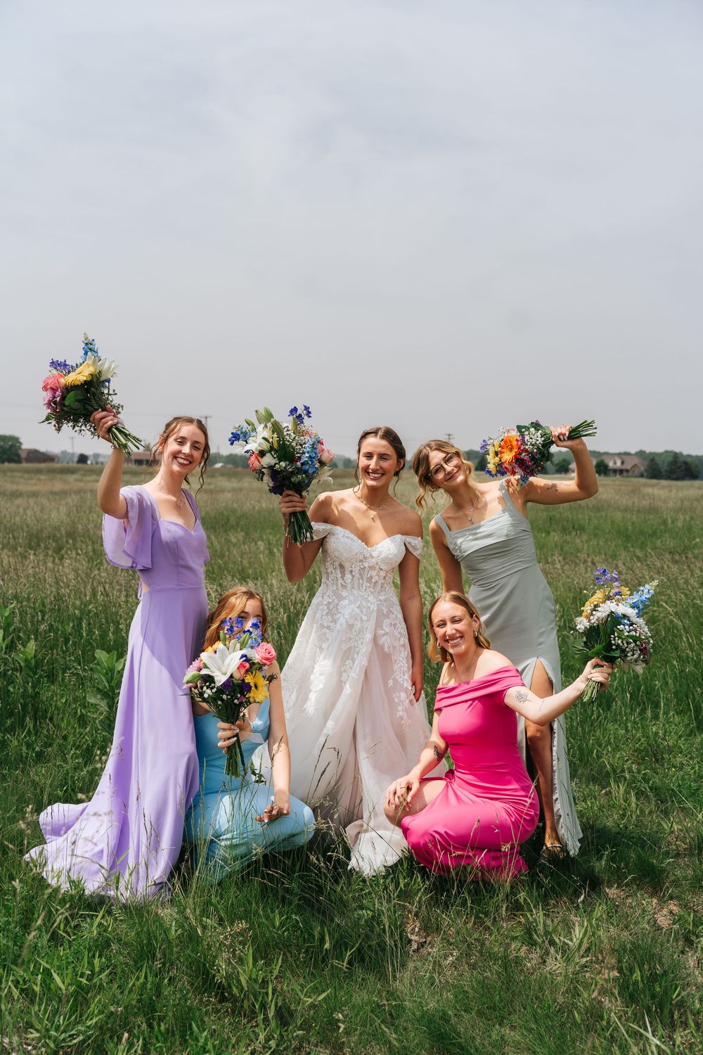 A wedding party in colorful dresses poses playfully in a green meadow with vibrant floral bouquets.