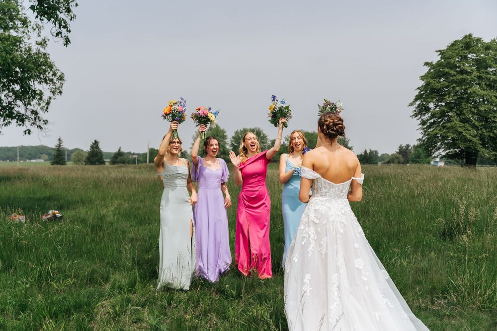 Wedding party members stand together outdoors holding vibrant floral bouquets against a natural landscape.