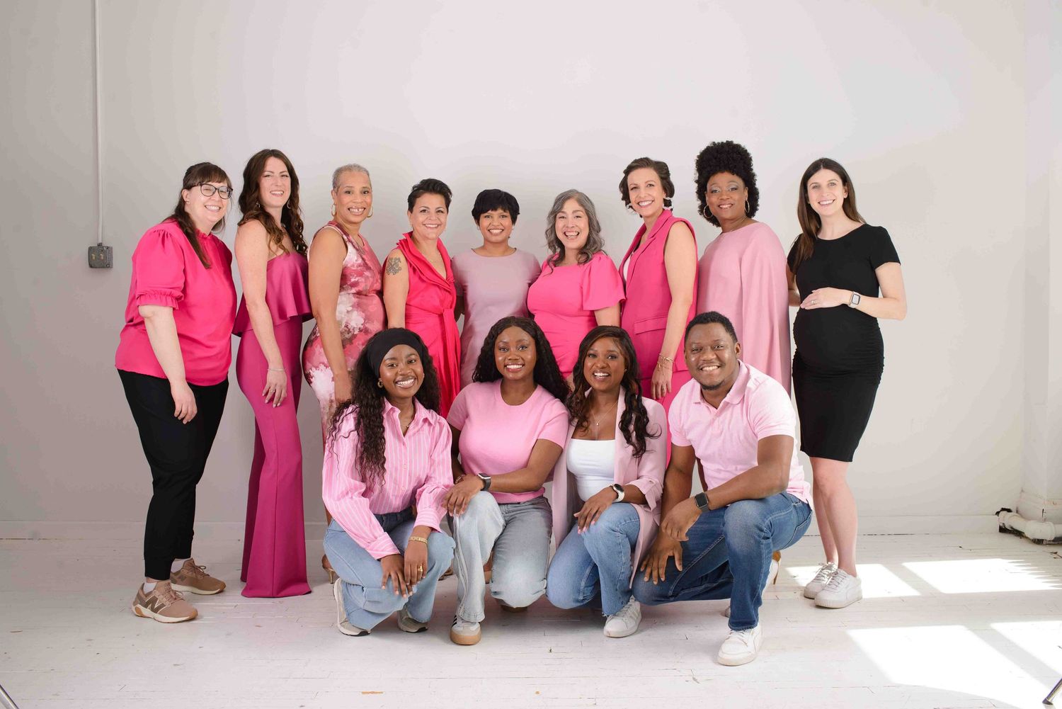 Group of people wearing pink and black clothing pose together in a white studio setting for breast cancer awareness.