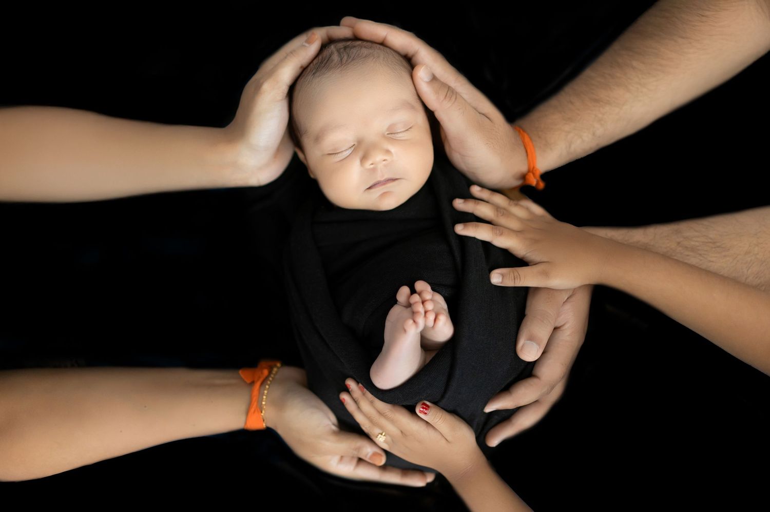 A series of hands holding and cradling a sleeping newborn baby against a black background.