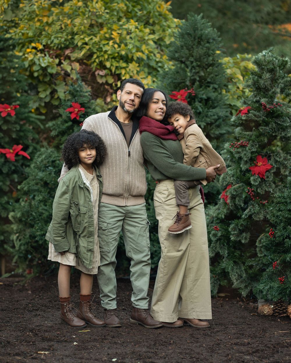 Family poses together in matching earth-toned outfits against a backdrop of green foliage and red poinsettias.
